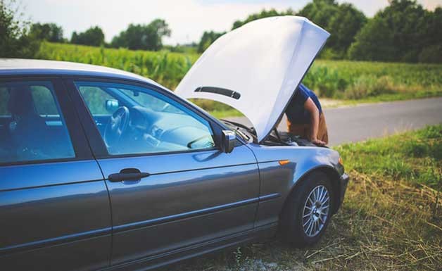 A man stands at the open hood of a car near a country road on a summer day