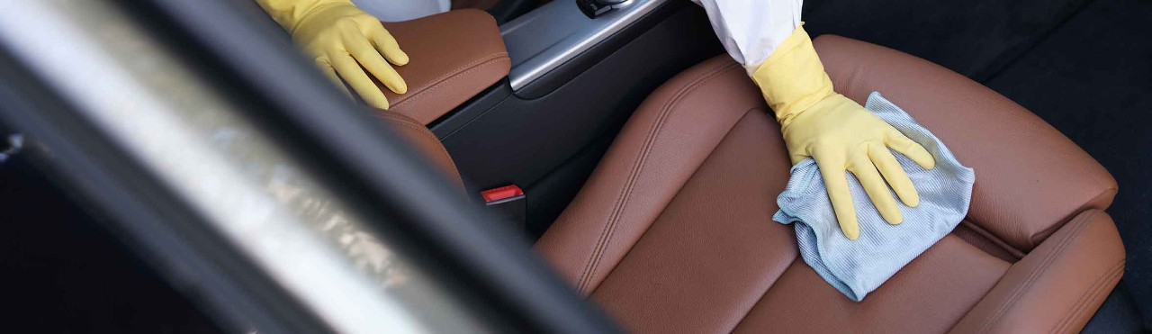 Man in protective suit and rubber gloves wiping leather car seat with rag with disinfectant closeup
