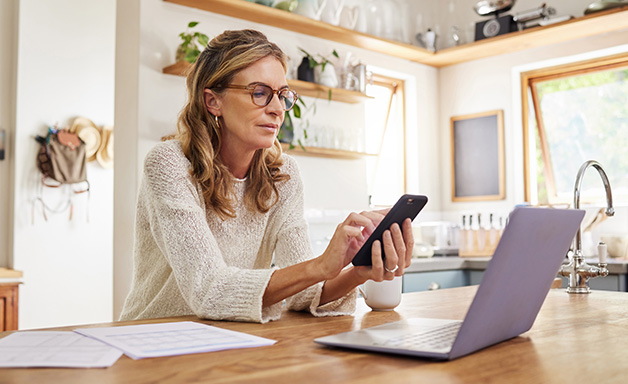 Shot of a mature woman typing on mobile phone while using a laptop at home